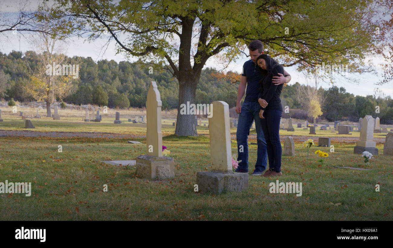 Sad boyfriend and girlfriend hugging at grave in lonely cemetery Stock Photo