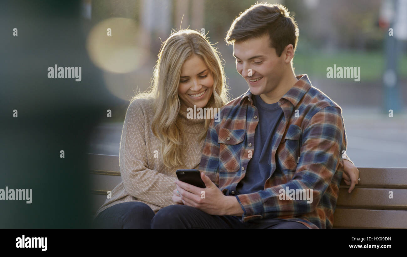 Selective focus view of smiling boyfriend and girlfriend sharing smartphone and sitting on bench Stock Photo