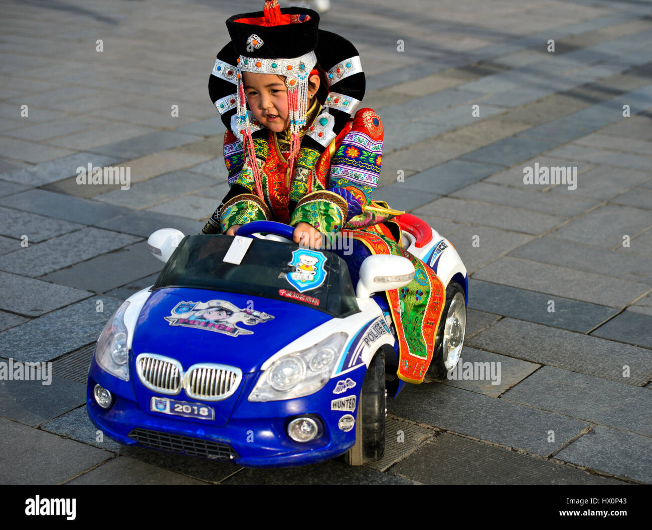Girl in traditional Deel clothing driving a toy car, police car, Festival of the Mongolian national traditional costume Stock Photo