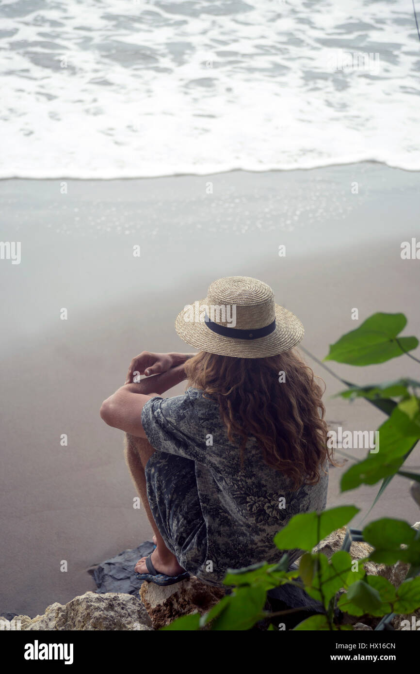 Indonesia, Java, woman wearing straw hat sitting on rock on the beach ...