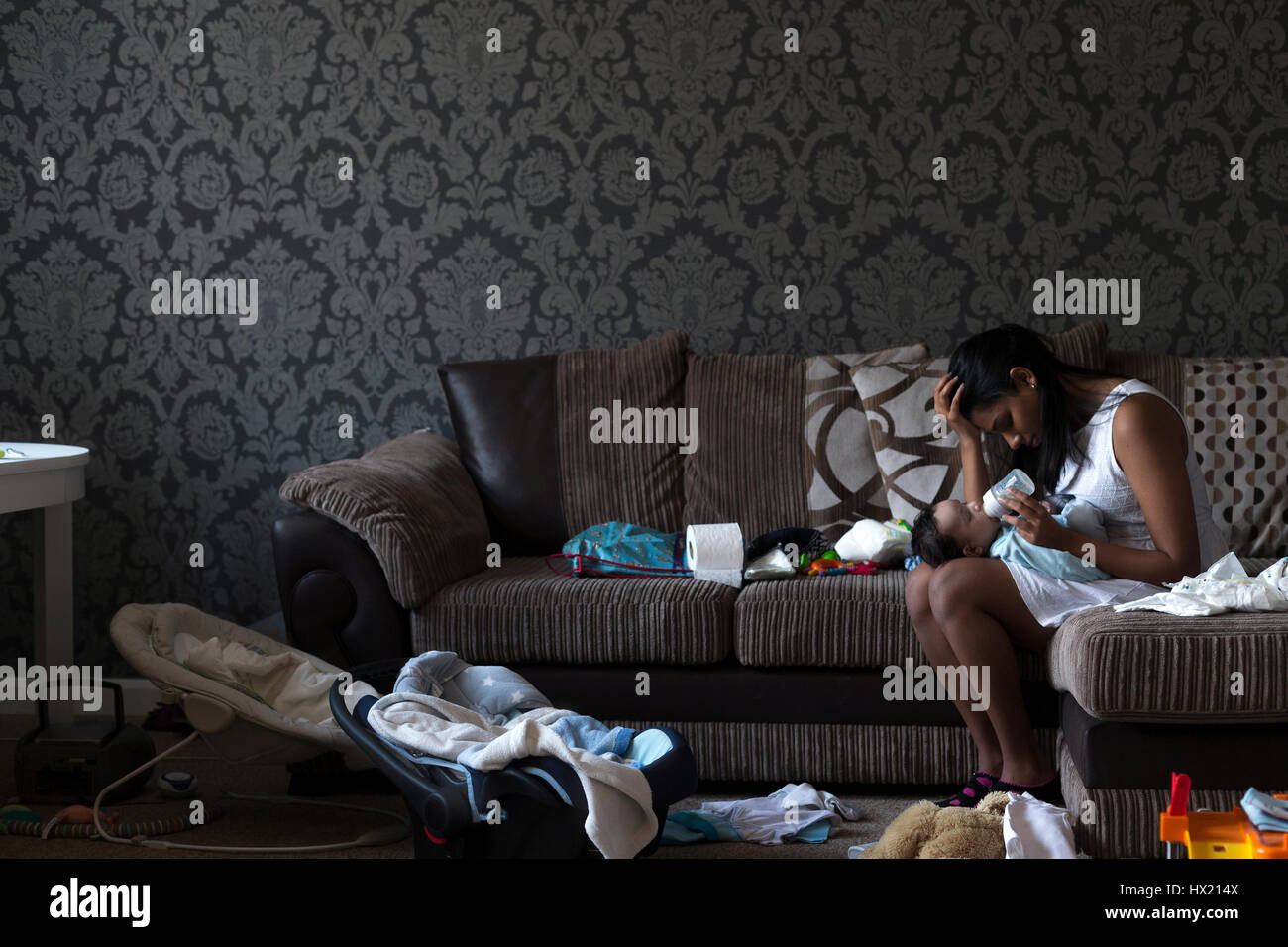 Stressed young mother sitting on her sofa whilst feeding her baby son. She has her head in her hand and is surrounded by mess. Stock Photo