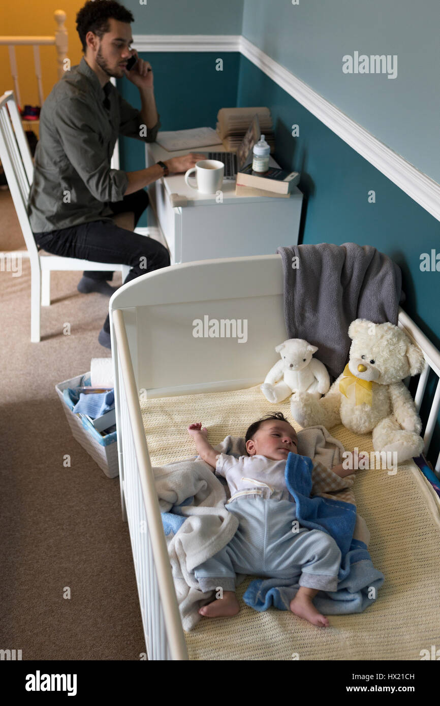 Young father working from home whilst his baby son is sleeping in his cot. Stock Photo
