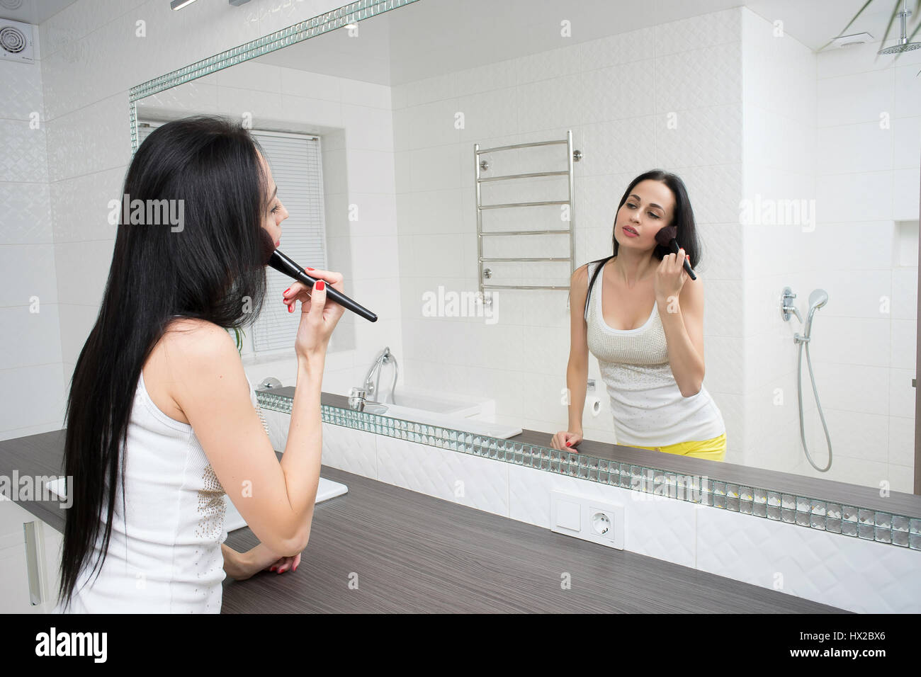 A young woman is standing in front of the bathroom mirror and putting on makeup. Stock Photo