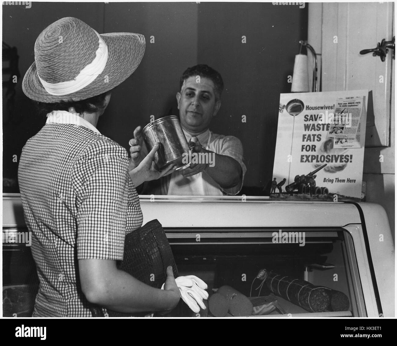 A housewife wearing a straw hat and white gloves reaches over the glass counter to hand a butcher a can of solid fat in exchange for money, after which it will be processed into ammunition for America's World War II efforts, 1942. Stock Photo