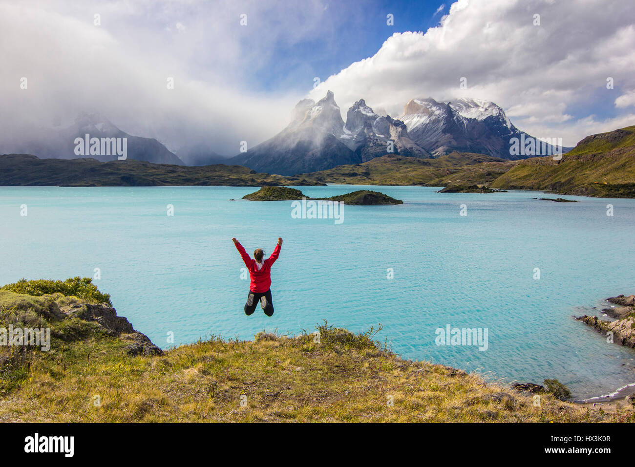 girl in red jacket with hands up jumping above lake in mountains Stock Photo