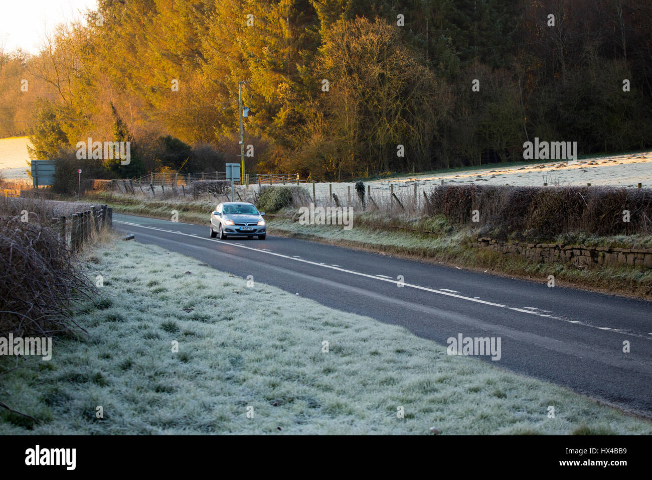 Vehicle travelling along a quiet road surrounded by frozen fields and the sunrise hitting the trees in the distance in Flintshire near to the town of Mold Stock Photo