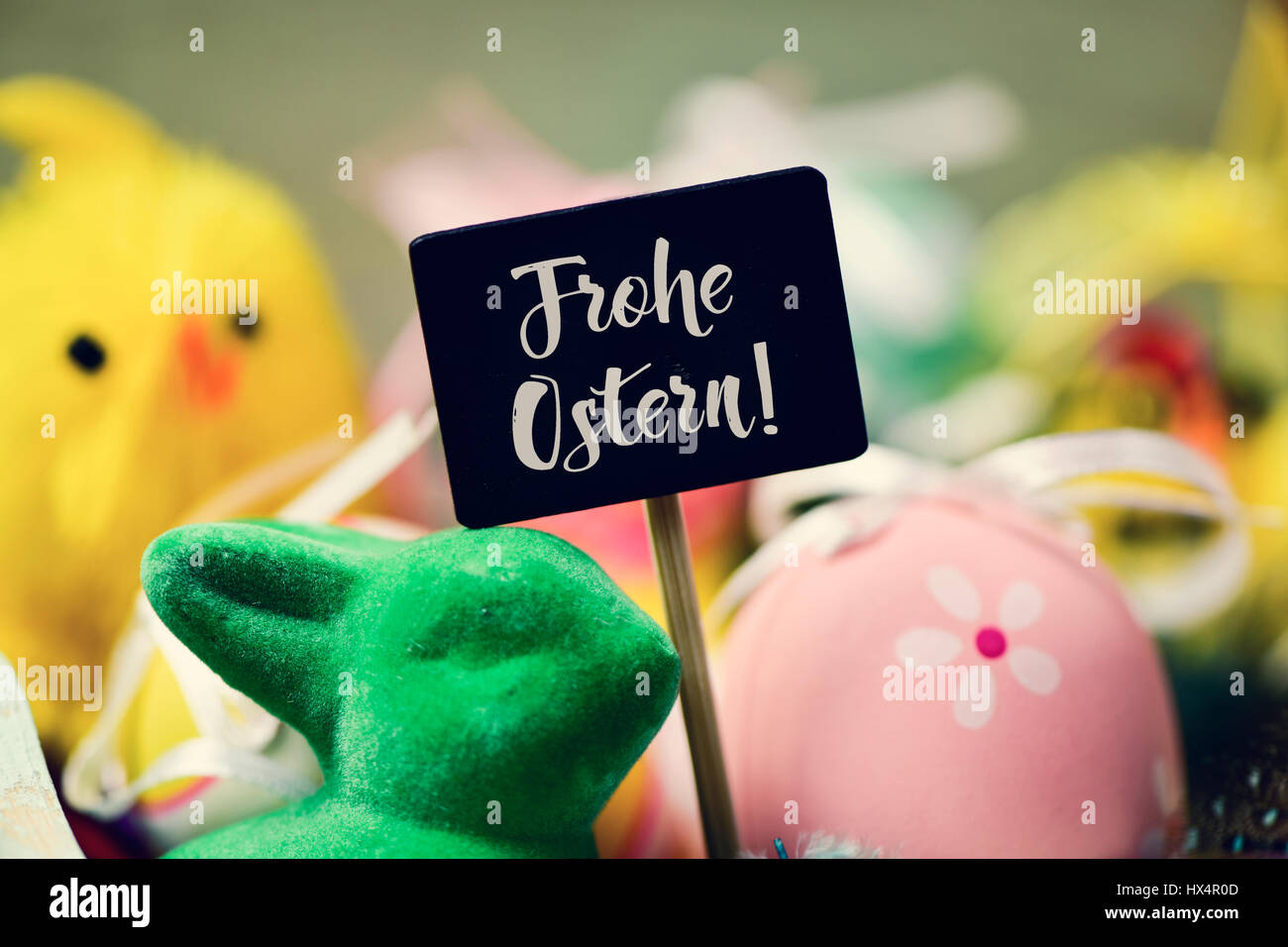 a chalkboard with the text frohe ostern, happy easter in german written in it and a pile of decorated easter eggs of different colors, a green rabbit  Stock Photo