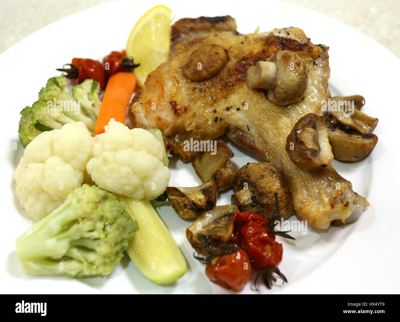 Oven grilled chicken leg and mushrooms, served with steamed cauliflower, courgette,carrot, cherry tomatoes and romanesco broccoli. Stock Photo