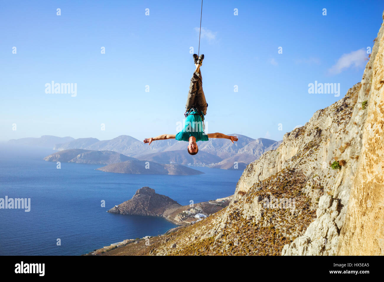 Cheerful rock climber swinging on rope upside down while fooling around, against view of coast Stock Photo