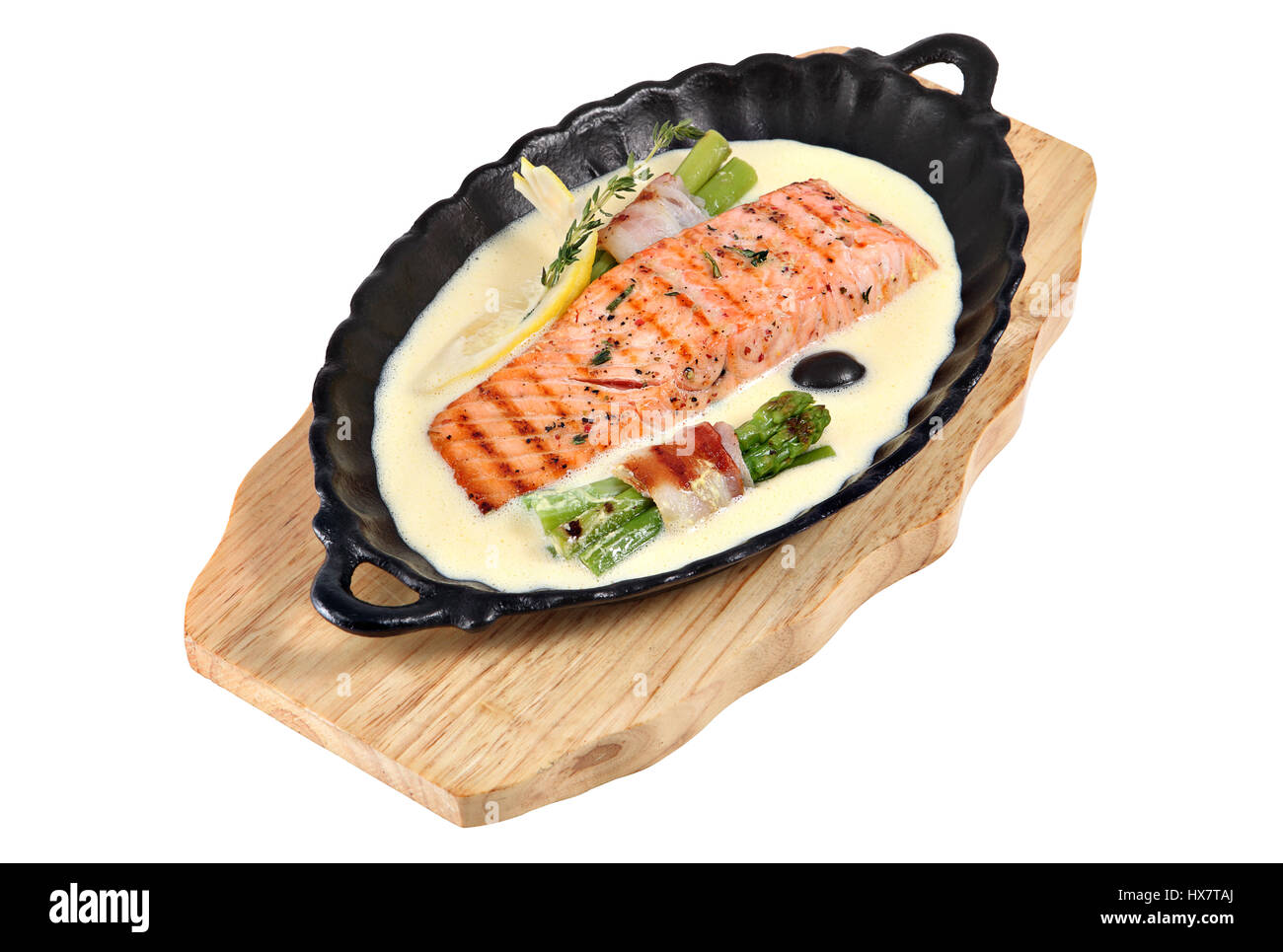 Roasted salmon in white sauce and Grilled green asparagus wrapped with bacon in cast iron oval the serving bowl on wooden plate, isolated on white bac Stock Photo