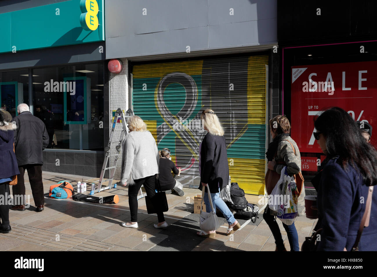 People walk past a street artist spray painting art on a closed shop shutter in Sheffield city centre England UK Stock Photo