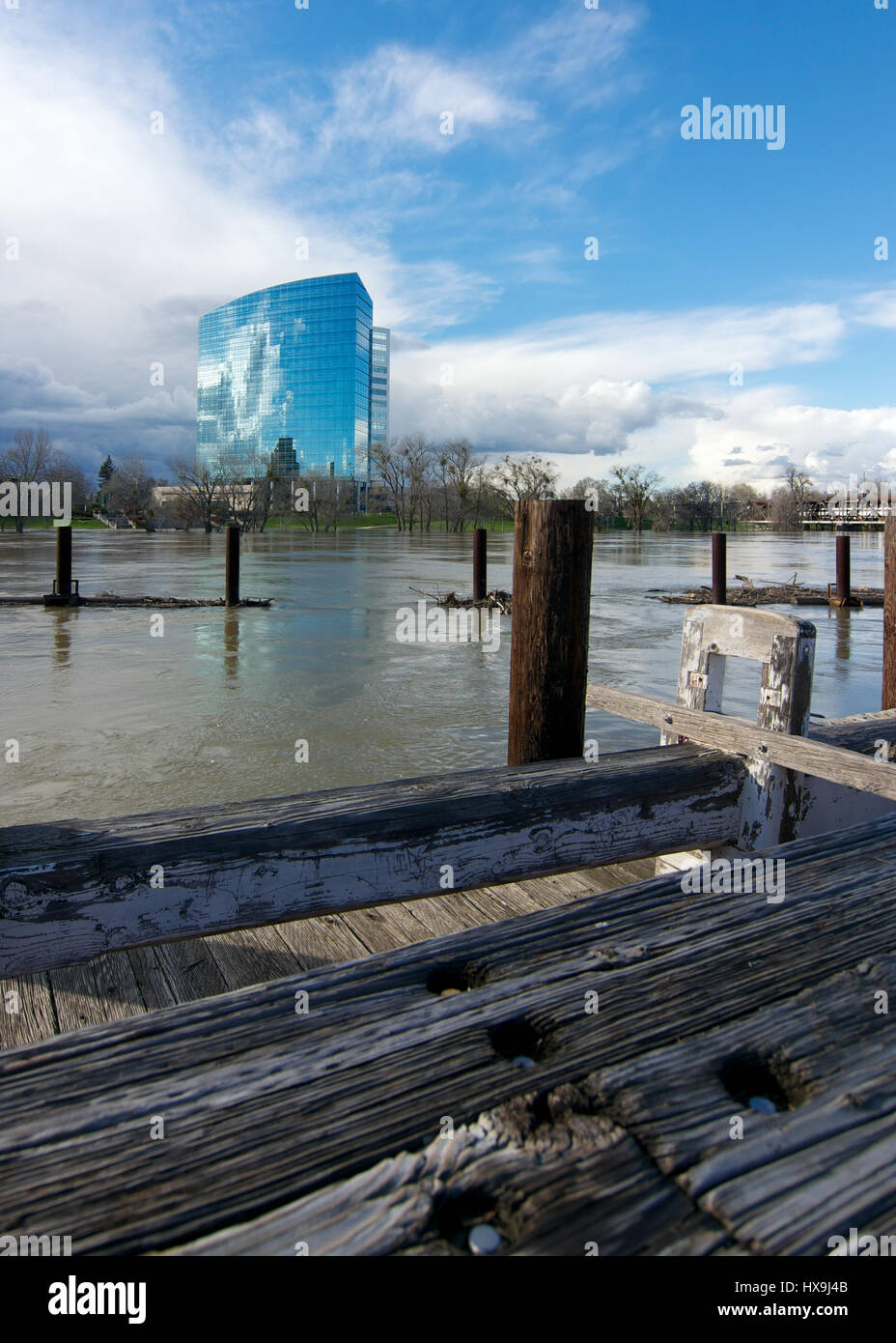 The mirror tower building in West Sacramento viewed from Old Sacramento Historic park on a partly cloudy day, featuring a full Sacramento River inbetw Stock Photo