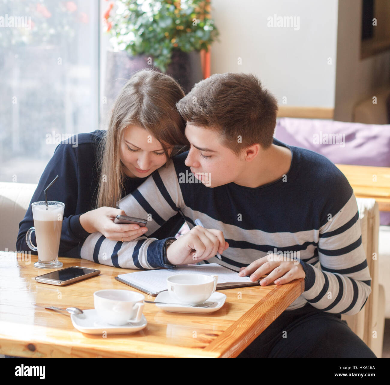 Beautiful young couple sitting in cozy coffee shop. Couple in love making planning of life and writing in notepad from smartphone. Stock Photo