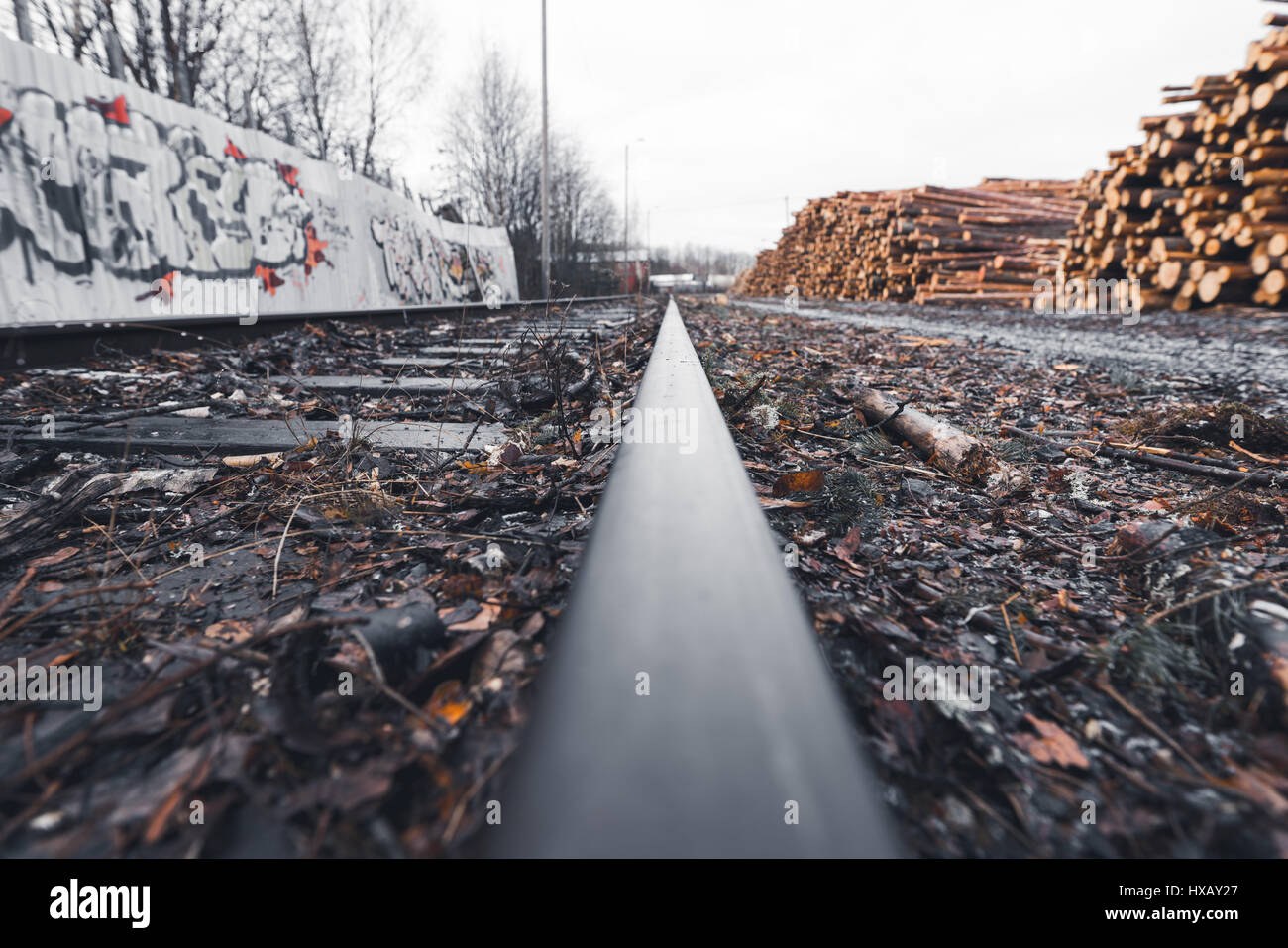 Wide and low angle view of railroad track covered by debris dividing blurred metal wall and pile of wood on overcast and wet day Stock Photo