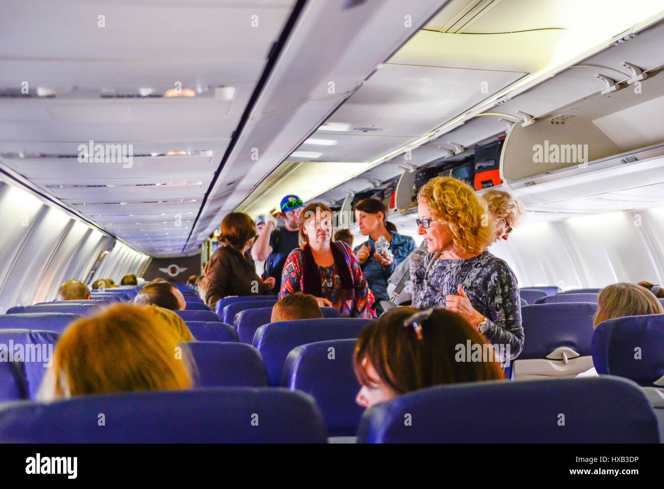 A view from the back of a commercial airplane of the cabin with people boarding, sitting & using overhead bins while flight attendants assist Stock Photo
