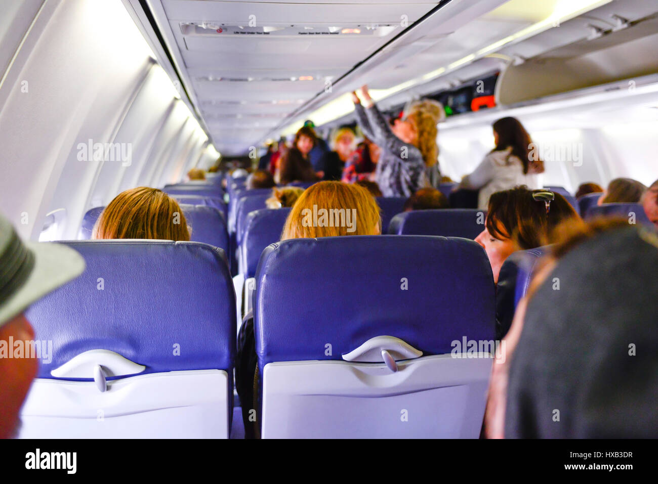 A view from the back of a commerical airplane of the cabin with people boarding, sitting & using overhead bins while flight attendants look on Stock Photo