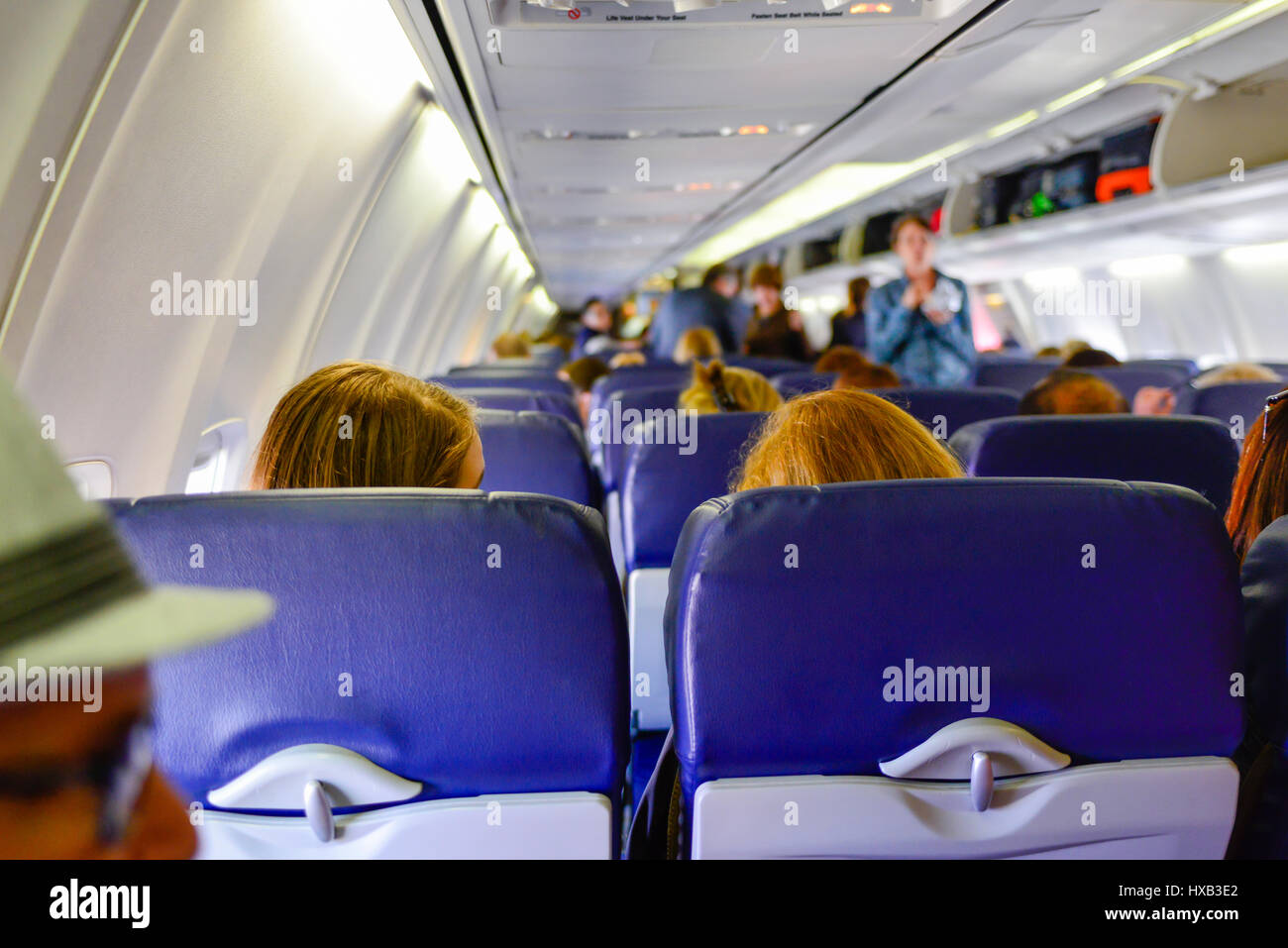 A view from the back of a commercial airplane of the cabin with people boarding, sitting & using overhead bins while flight attendants look on Stock Photo