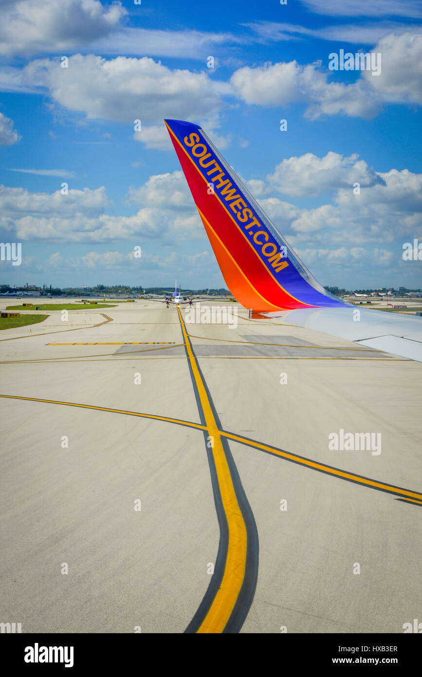 A wing tip of a Southwest Airlines airplane taxis with yellow paint markings on tarmac crossing lines with blue skies and white clouds Stock Photo