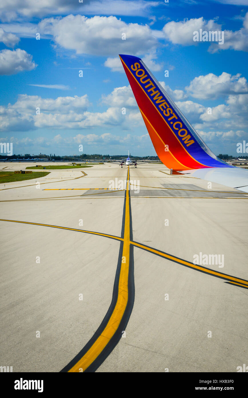 A wing tip of a Southwest Airlines airplane taxis with yellow paint markings on tarmac crossing lines with blue skies and white clouds Stock Photo