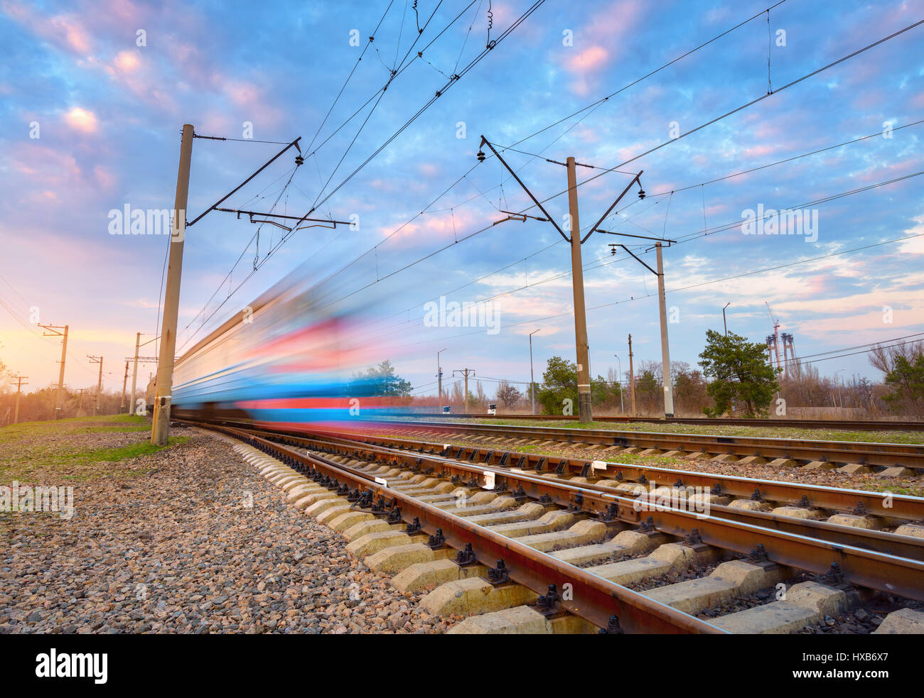 High speed blue passenger train in motion on railroad at sunset. Blurred commuter train. Railway station against colorful sky. Railroad travel, railwa Stock Photo