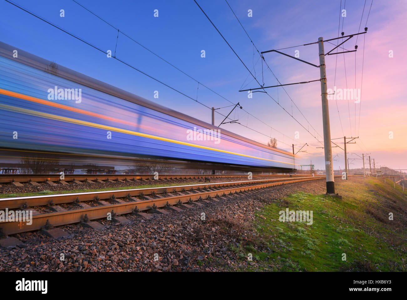 High speed passenger train in motion on railroad at sunset. Blurred commuter train. Railway station against colorful blue sky. Railroad travel, railwa Stock Photo