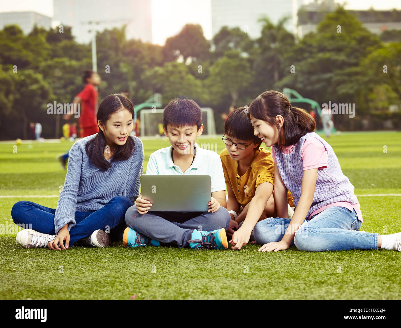 four asian elementary school boys and girls sitting on playground using laptop computer together. Stock Photo