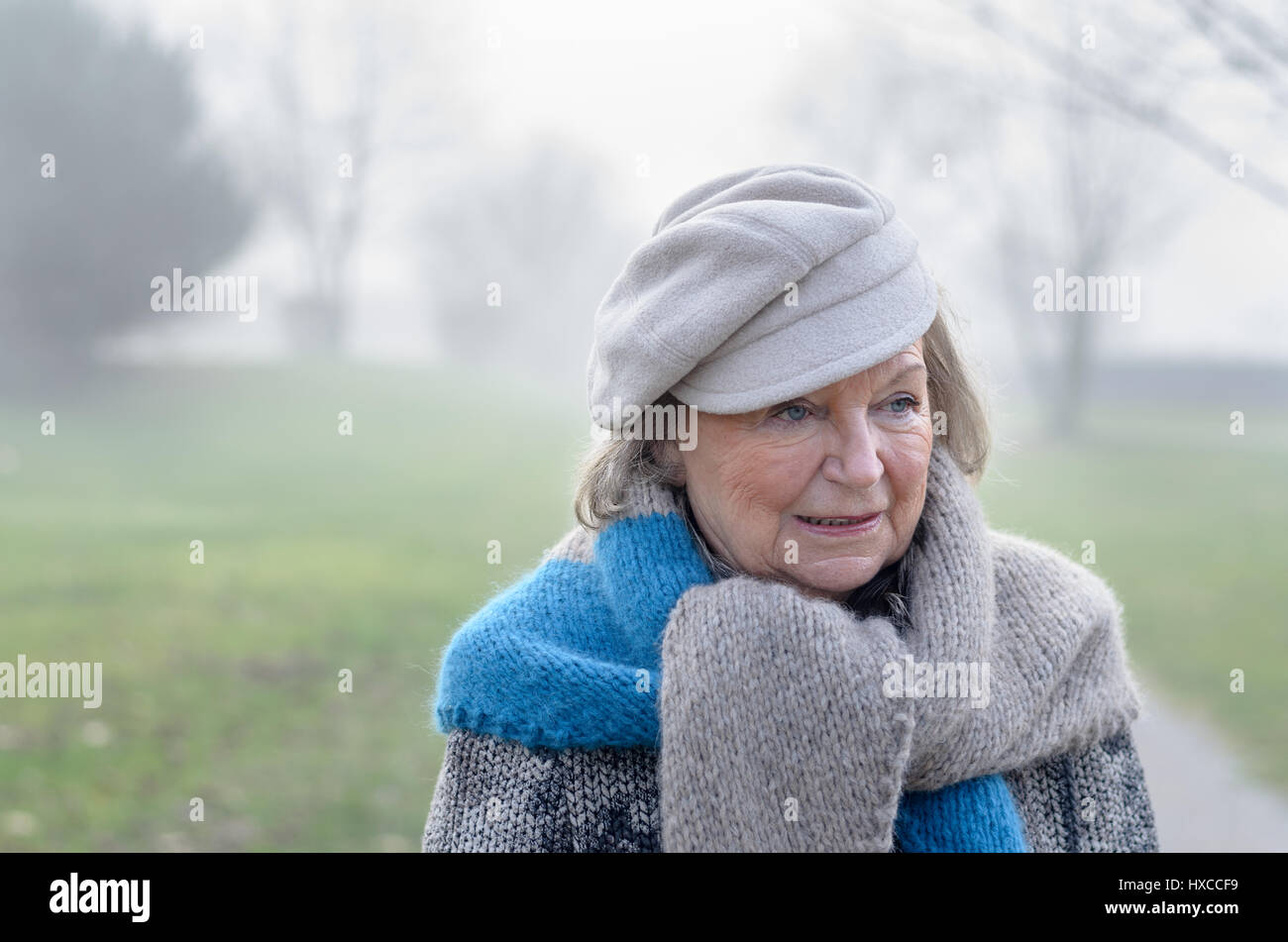 Stylish elderly woman wearing a grey scarf and beret walking on a rural road on a cold misty winter day, close up head and shoulders Stock Photo