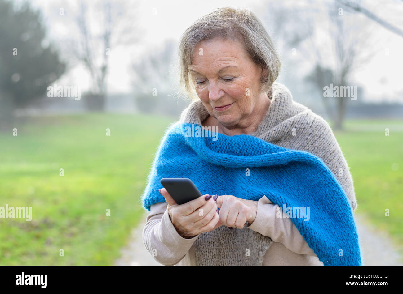 Attractive tech savvy elderly woman using a mobile phone texting a message to a friend or browsing for a number to call, on a rural lane on a misty wi Stock Photo