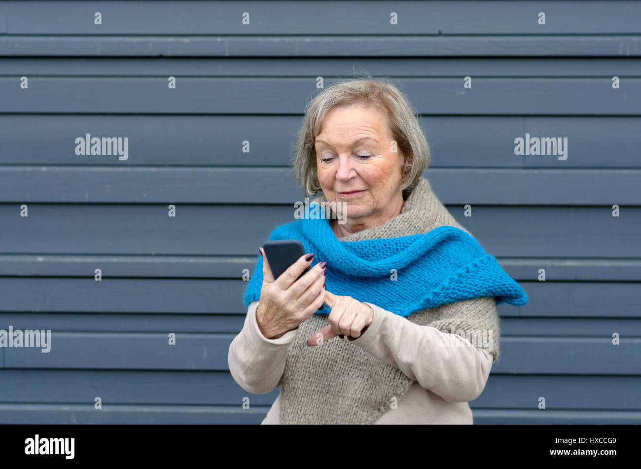 Attractive tech savvy elderly woman using a mobile phone texting a message to a friend or browsing for a number to call, grey metal background with co Stock Photo