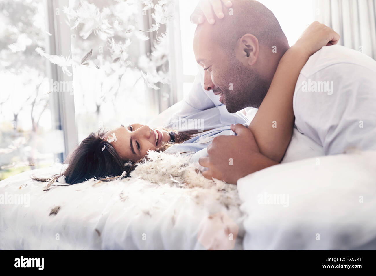 Pillow feathers falling around playful, affectionate couple on bed Stock Photo