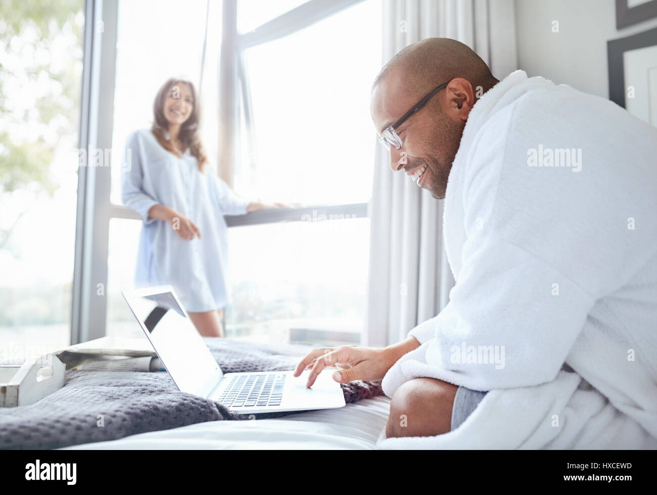Smiling woman watching boyfriend in bathrobe reading laptop on bed Stock Photo