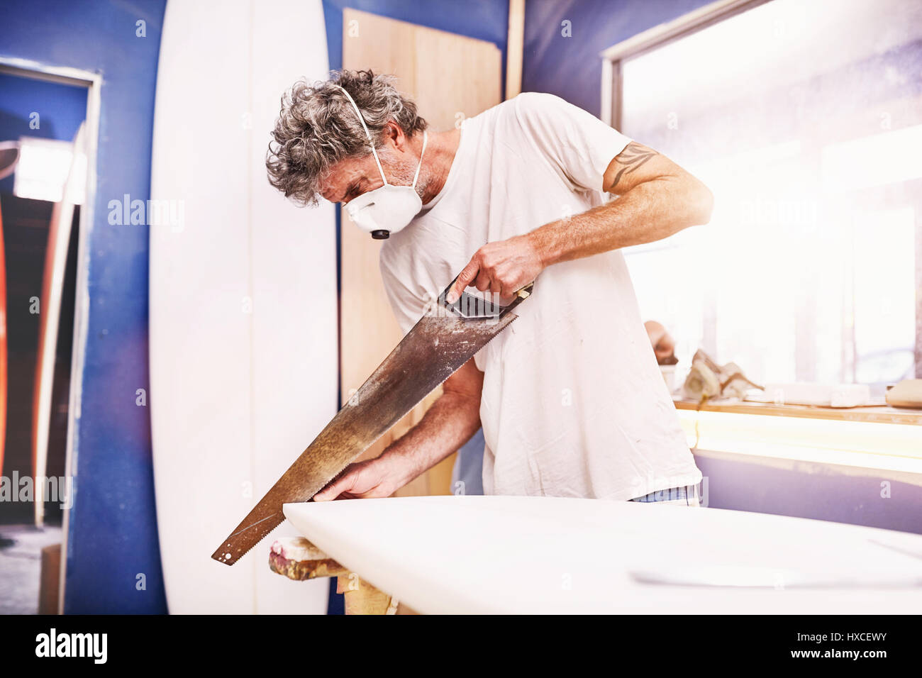 Male surfboard designer wearing protective mask and using saw in workshop Stock Photo