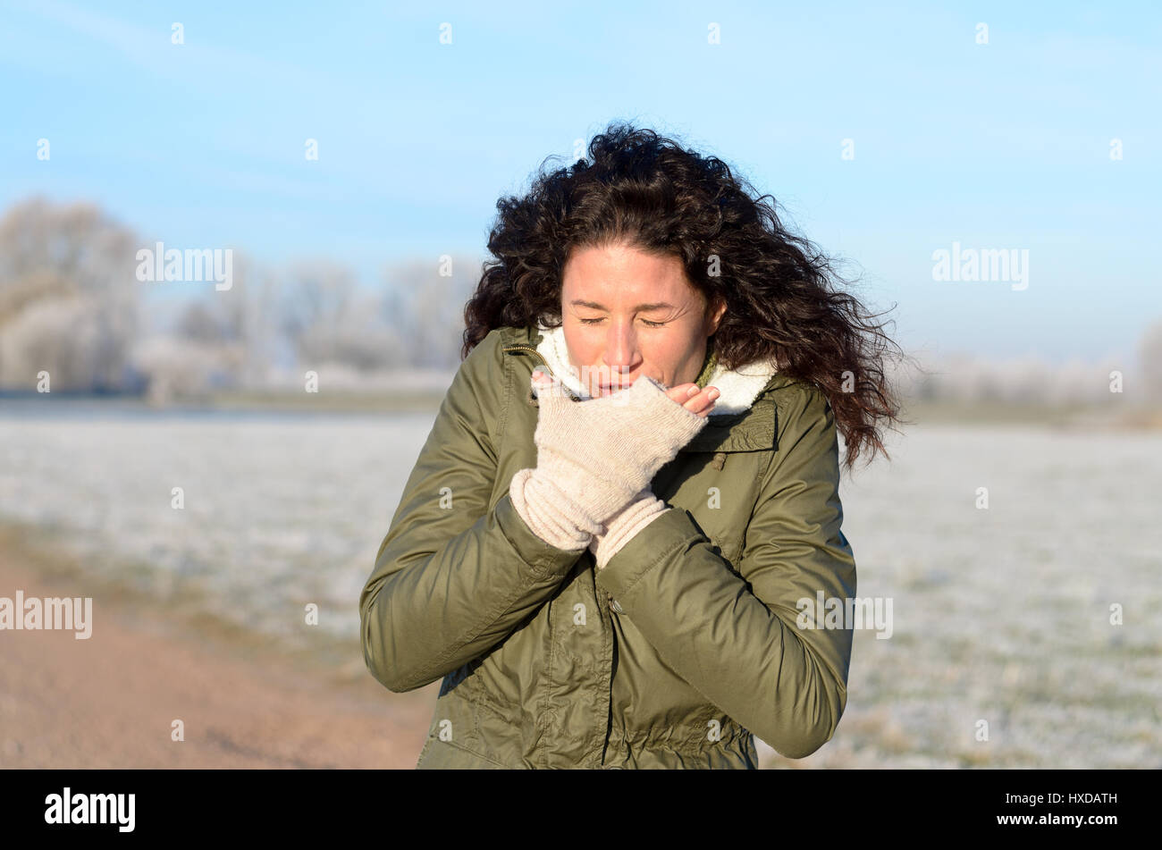 Young woman with a seasonal cold and flu coughing into her gloved hand as she stands on a rural road in a cold frosted winter landscape in sunshine, c Stock Photo