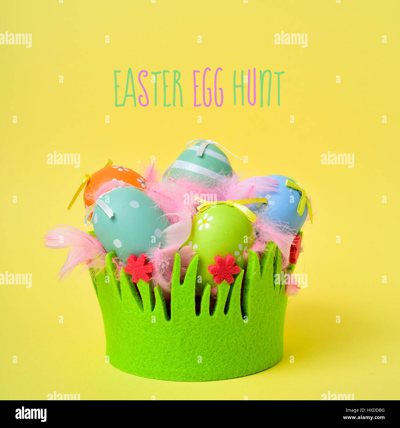 the text text easter egg hunt and a pile of different decorated easter eggs and pink feathers in a basket decorated as grass on a yellow background Stock Photo
