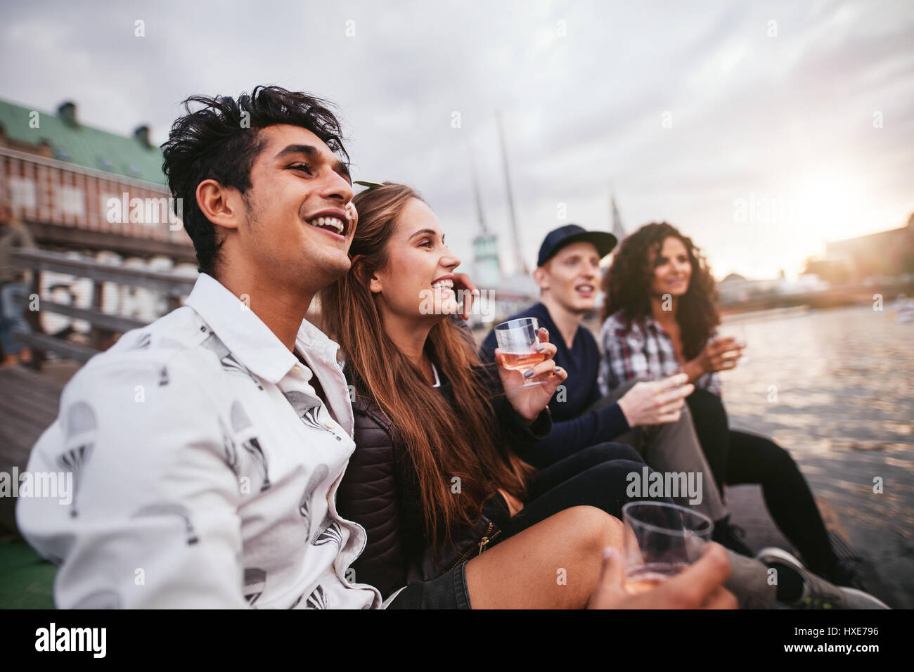 Group of young people having fun with drinks. They are sitting together by the lake and looking away smiling. Stock Photo
