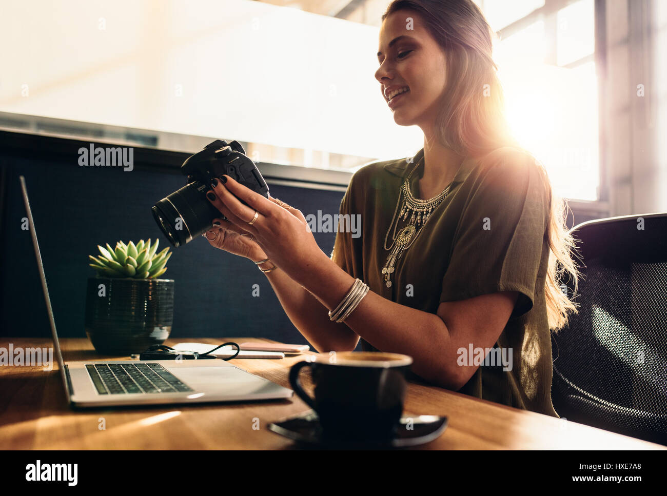 Young woman looking at camera while working on laptop. Young photographer with her camera and laptop on her desk. Stock Photo
