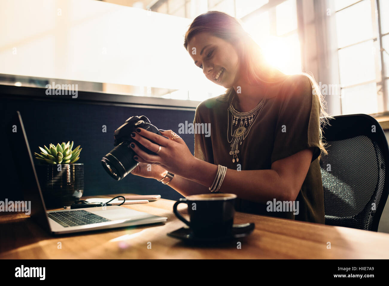 Young female vlogger looking at camera while working on laptop. Photographer with her camera and laptop on her desk. Stock Photo