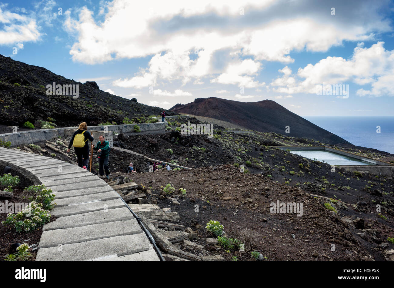 Cumbre Vieja, Fuencaliente, La Palma. . Some hikers taking the man made path that passes the water reservoir that is used to supply the water plantations.  It's a bright day with fast moving clouds along the coastline. Stock Photo