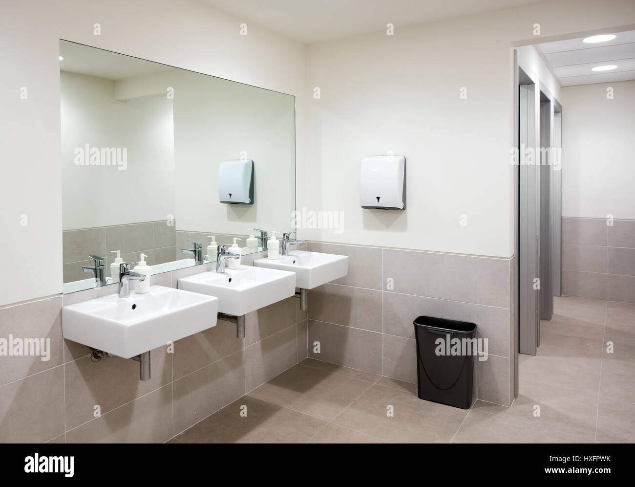 Public bathroom or restroom with a row of three hand basins with soap dispensers below a large mirror and neutral beige tiled decor Stock Photo