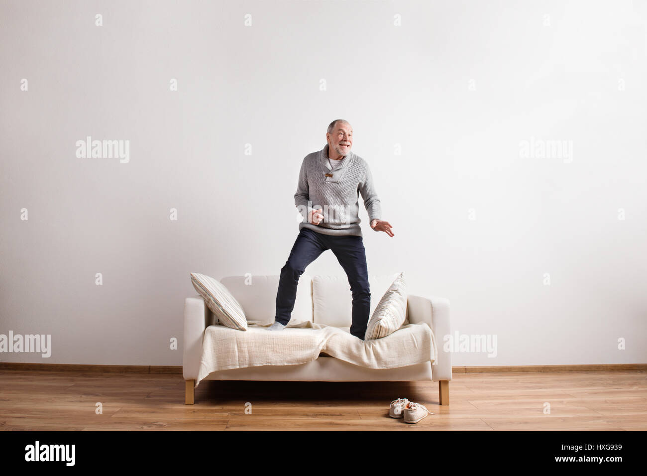 Handsome senior man standing on couch, dancing, having fun. Studio shot against white wall Stock