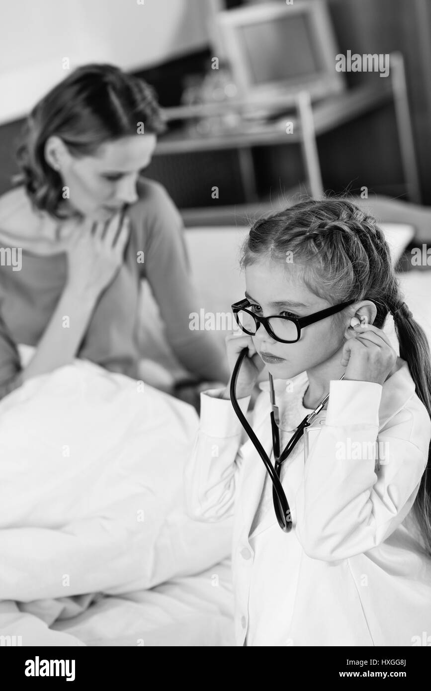 Black and white photo of little girl doctor with stethoscope standing near sick woman in hospital bed Stock Photo