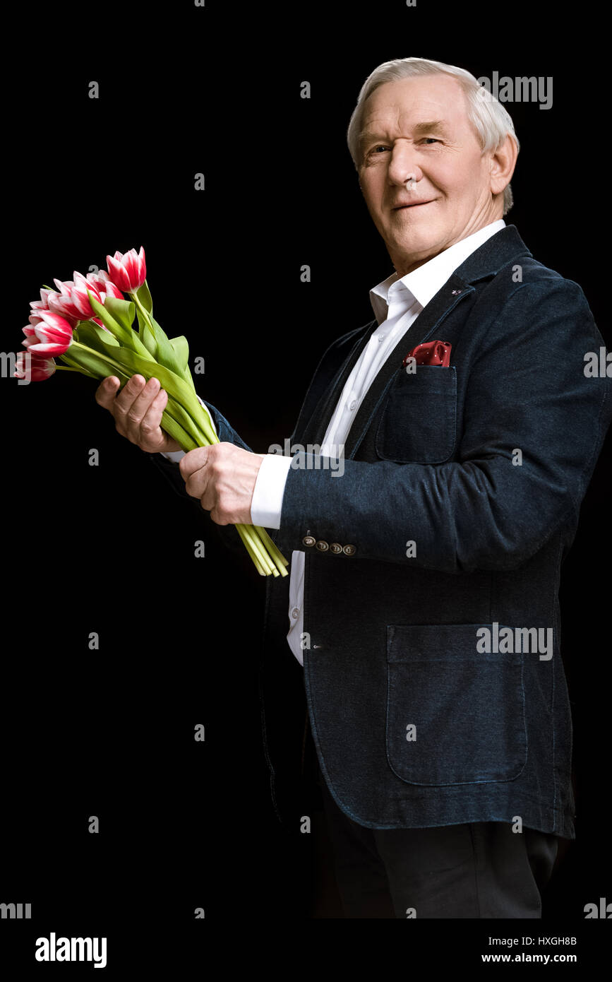 Smiling senior man in suit holding beautiful tulips bouquet on black, spring holiday concept Stock Photo