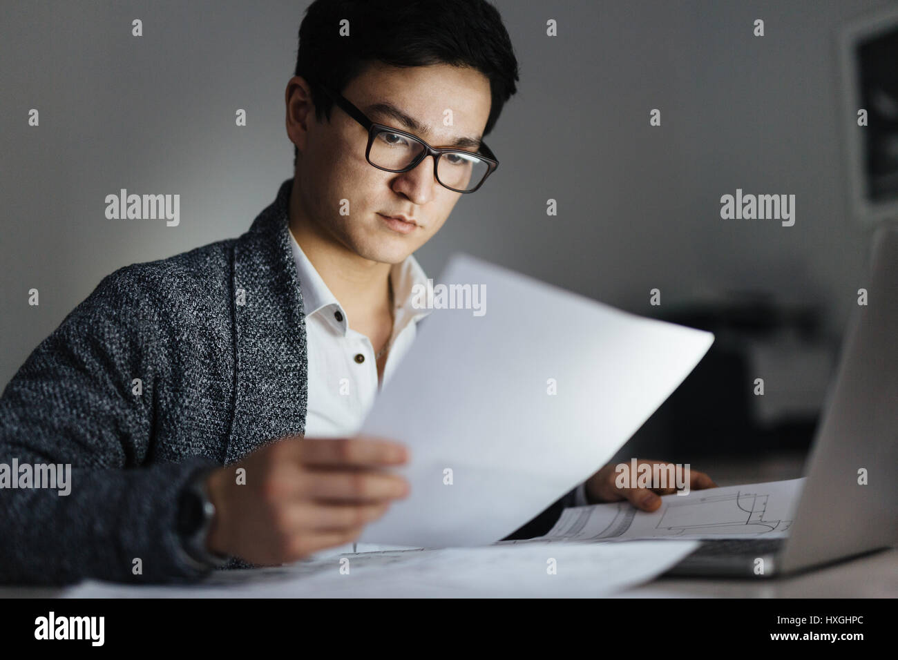 Portrait of busy Asian man wearing glasses and casual wear sorting documentation and working with laptop in dark room late at night, his face lit up b Stock Photo