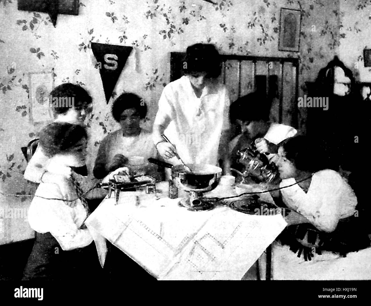 Illustration showing a group of excited housewives sitting around a suburban kitchen table and using a variety of electrical appliances, including a waffle iron, fondue pot, chafing dish and kettle, 1912. Stock Photo
