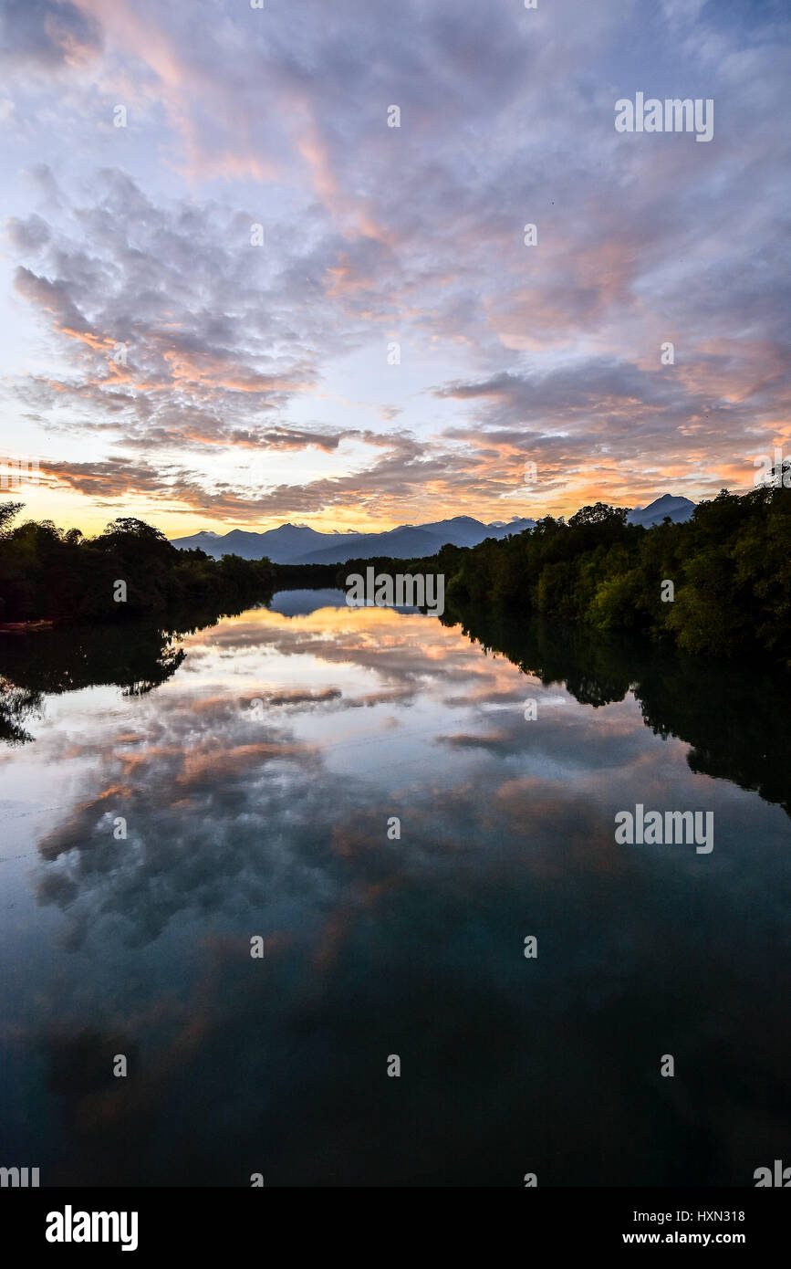clouds and sunset sky reflecting in wide mirror like still river with tree linded banks and montain range horizon. Stock Photo