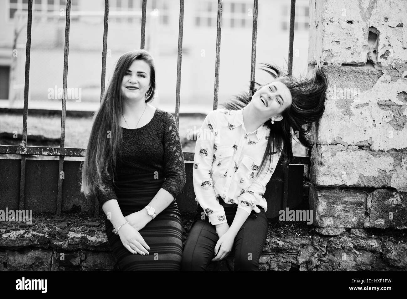 Two young teenages girl sitting against iron fence and moving with they hair. Plump girl vs thin. Friends teen. Stock Photo