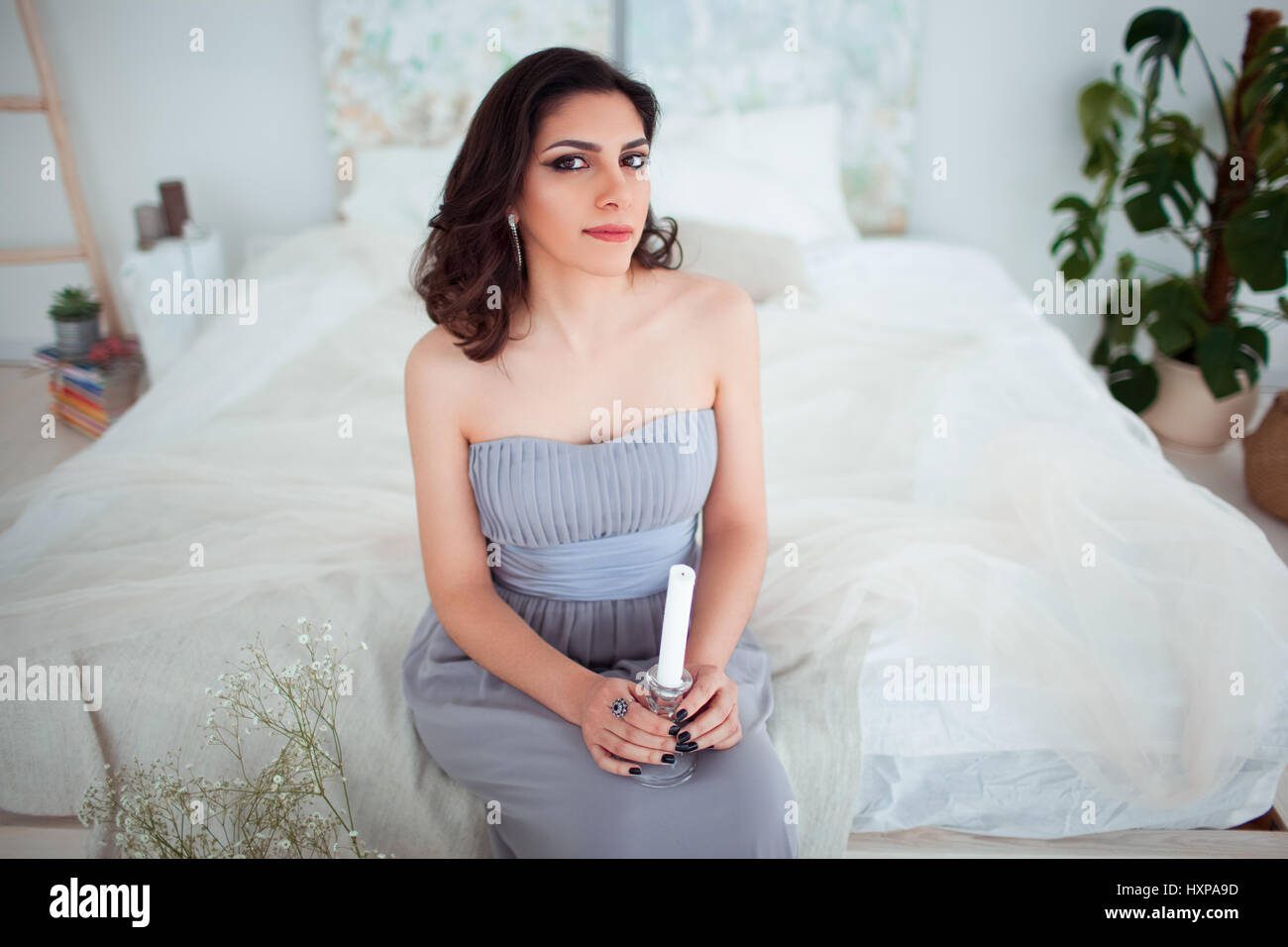 Cute girl with makeup in a blue evening dress sitting on the bed. In the hands holding a candle. Stock Photo