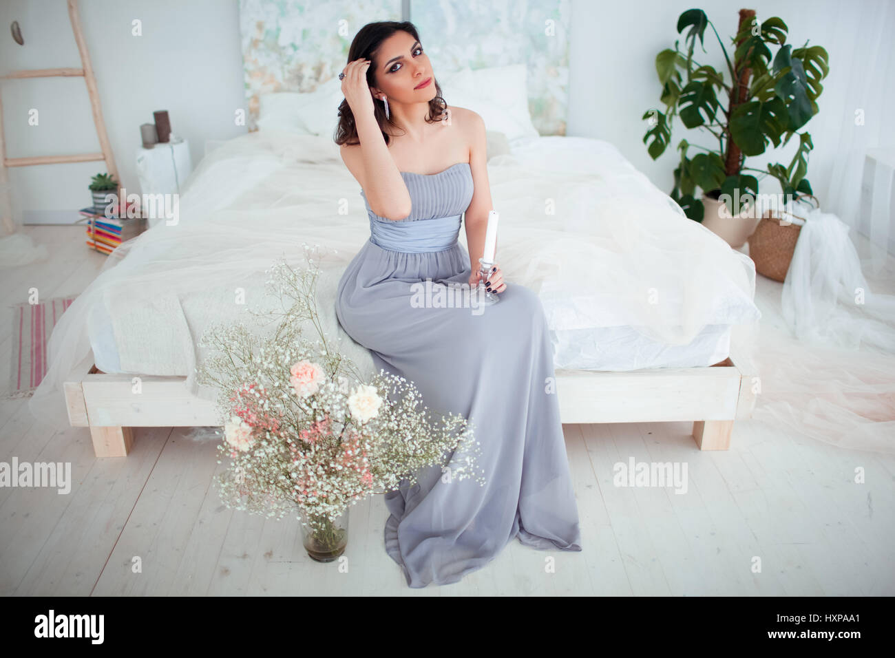 Cute girl with makeup in a blue evening dress sitting on the bed. In the hands holding a candle. Stock Photo