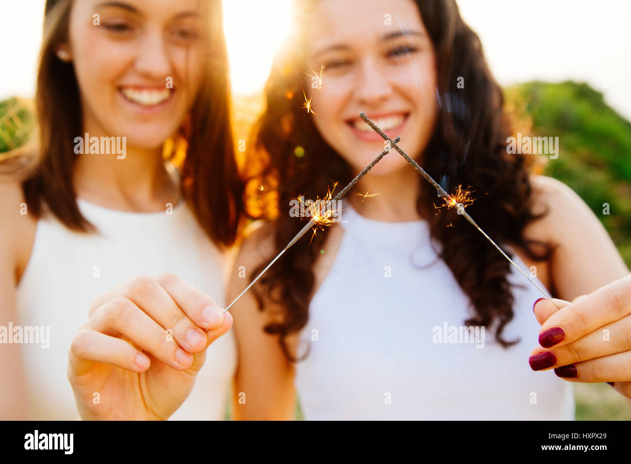 Happy female friends holding sparklers outdoors at sunset. Focus on sparks. Stock Photo