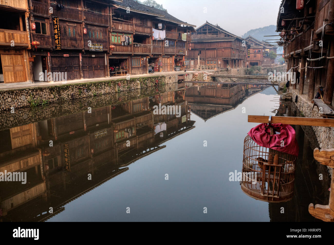 Zhaoxing Dong Village, Guizhou Province, China -  April 9, 2010: Wooden houses of farmers reflected in the water of the rural river, cage with a bird  Stock Photo